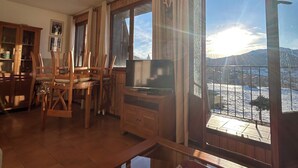 Un salón acogedor con una mesa de comedor de madera, sillas, un televisor de pantalla plana sobre un mueble de madera y un ventanal que ofrece vistas de un paisaje nevado.