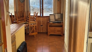 Una habitación acogedora con paredes de madera, suelo de baldosas, una zona de comedor con sillas, un televisor y vistas a montañas nevadas.