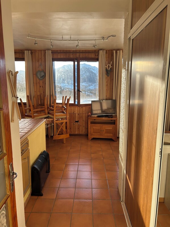 Une pièce chaleureuse aux murs en bois, dotée d’un sol carrelé, d’un coin repas avec des chaises, d’un téléviseur et offrant une vue sur des montagnes enneigées.