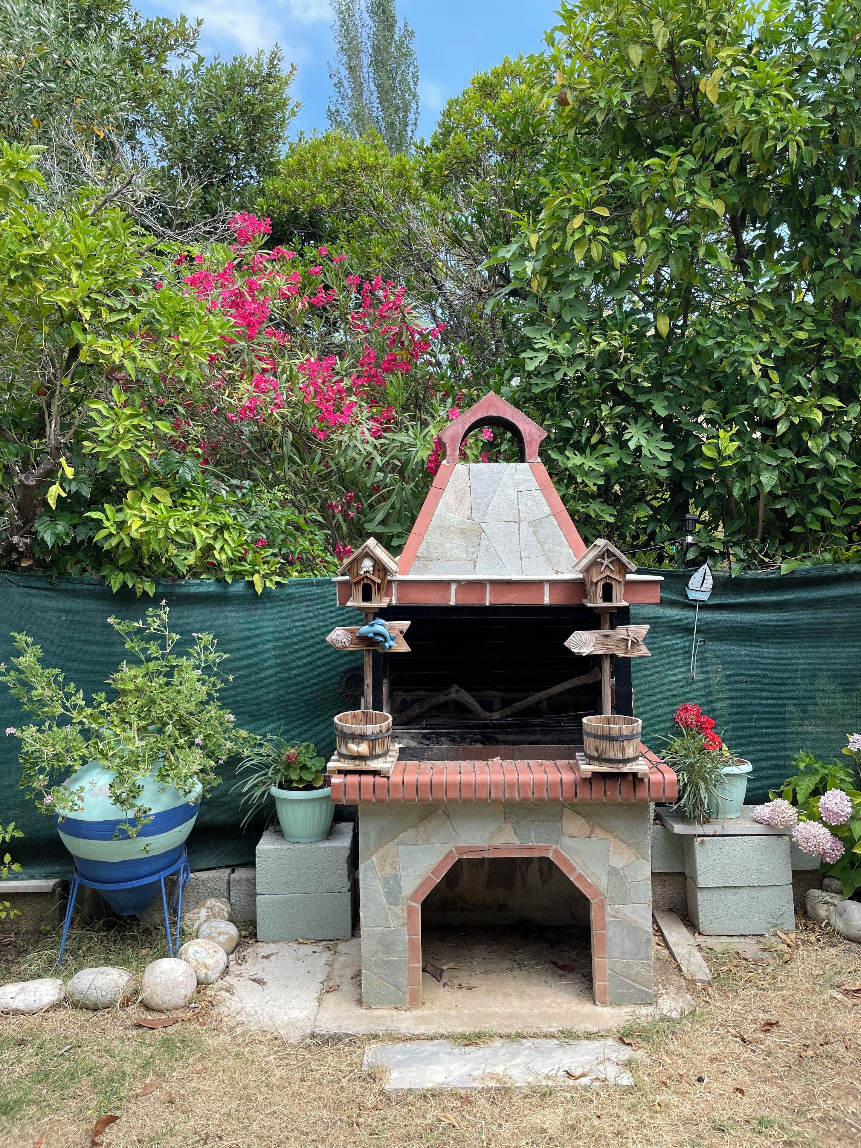 A stone-built outdoor oven with a tiled roof, surrounded by potted plants and a green fence.