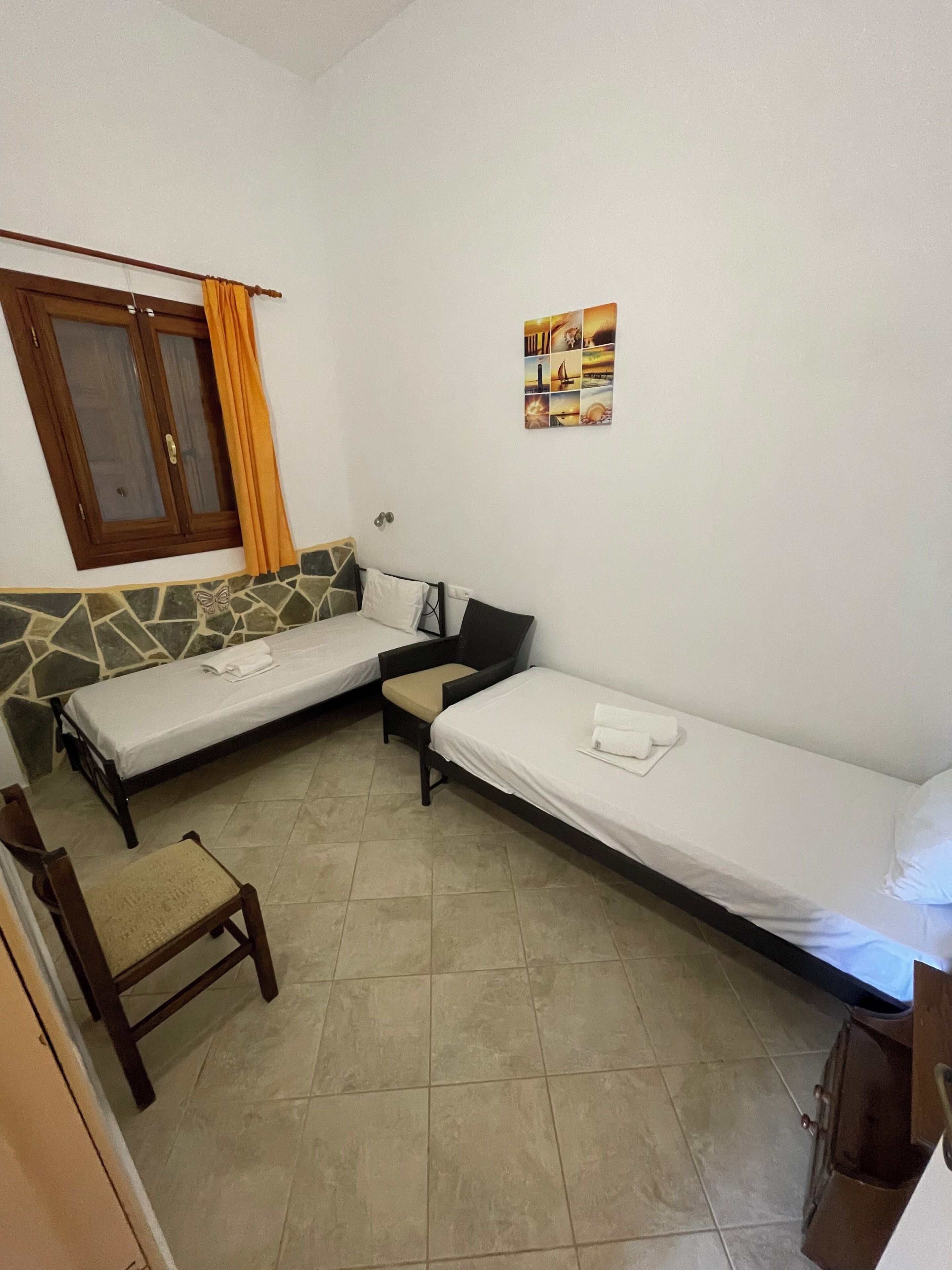 A small, well-lit room with two single beds, a chair, and a window with curtains.