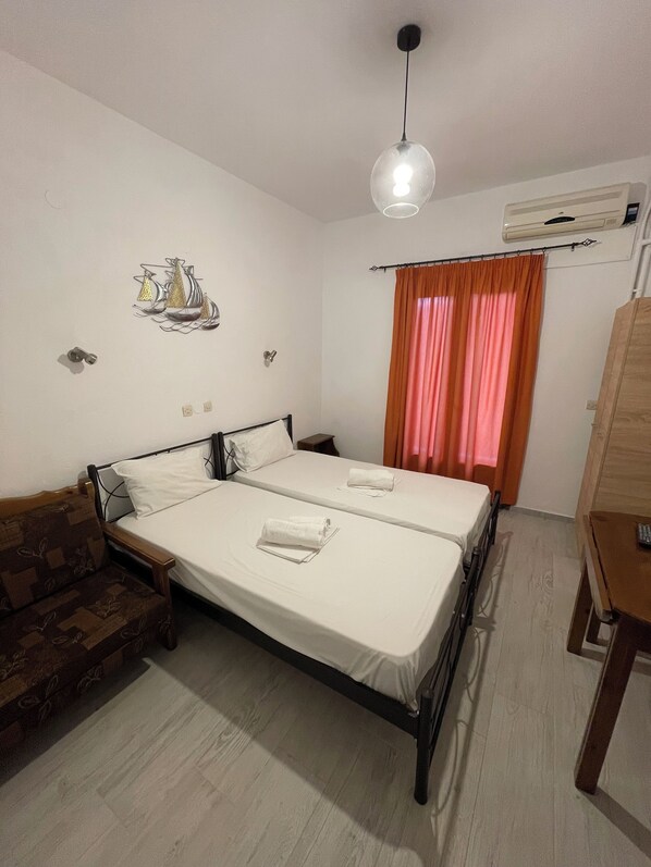 A double bed with white linens, a wooden nightstand, a small table with a remote, a decorative wall hanging, and an air conditioning unit. - Delfinia Studios (South Pelion)