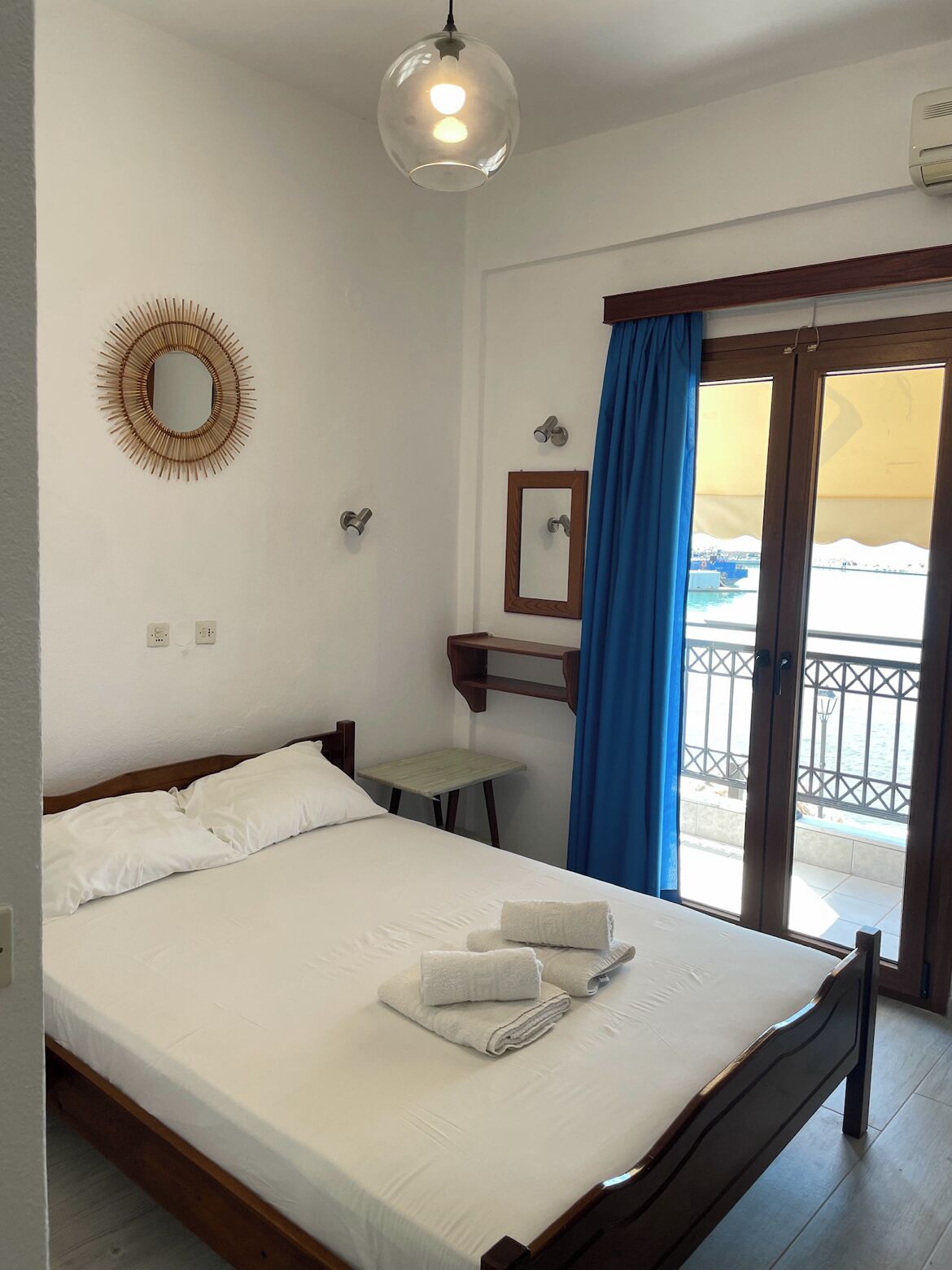 Hotelkamer met een bed, een klein tafeltje, een spiegel en uitzicht op zee vanaf een balkon met blauwe gordijnen.