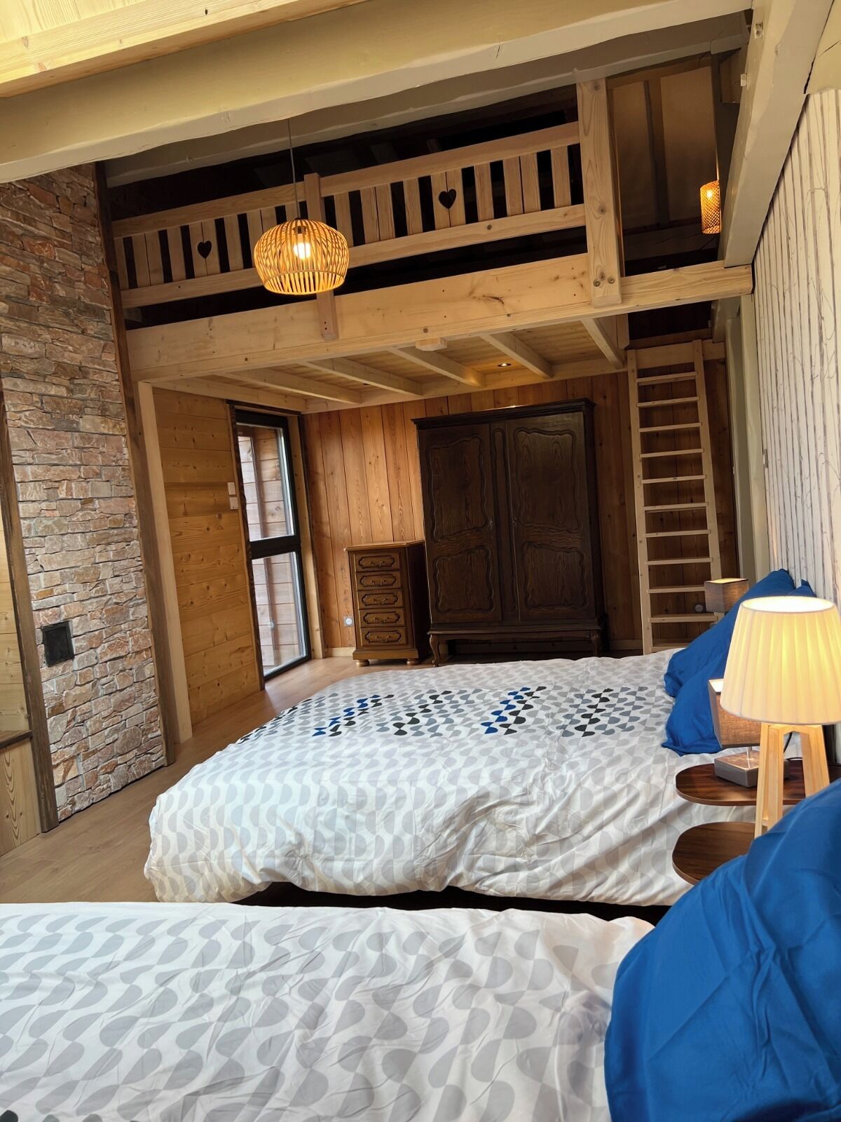 A loft-style bedroom with a high ceiling, exposed wooden beams, a large bed with patterned bedding, a wooden wardrobe, and a hanging light fixture.