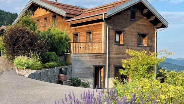 Una casa de madera de dos pisos con techo inclinado, rodeada por un jardín ajardinado con lavanda y otra vegetación.