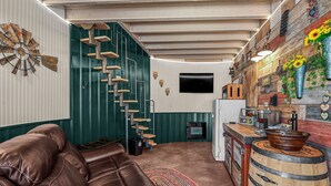 A cozy living room with a leather sofa, a spiral staircase, a flat-screen TV, and a rustic kitchen area with a wooden barrel.