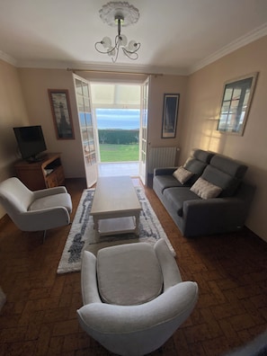 Een woonkamer met een bank, fauteuil, salontafel, televisie en uitzicht door een open deur.