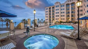 A hotel pool area with lounge chairs, a hot tub, and a building with balconies.