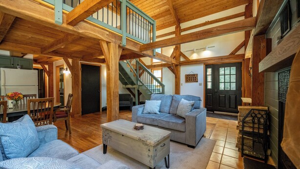A spacious living room with a high ceiling, wooden beams, a staircase, a fireplace, and a cozy seating area.