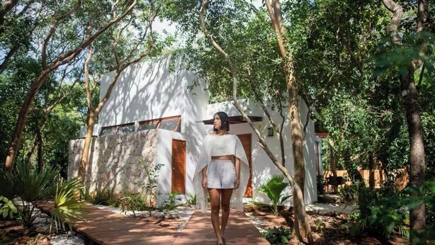 A person walking on a wooden pathway towards a white building with wooden doors and windows, surrounded by lush greenery. - Baalamkaab Flamenco Studio (Chichimilá)