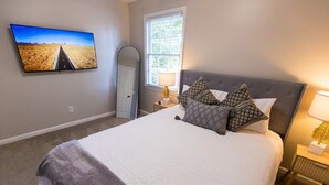 Een slaapkamer met een groot bed, een televisie aan de muur, een raam met uitzicht op groen, en een nachtkastje met een lamp.