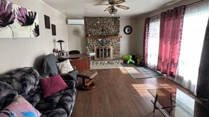 Ein Wohnzimmer mit Kamin aus Stein, Deckenventilator, einer Couch mit gemusterten Kissen und einem großen Fenster mit Vorhängen.
