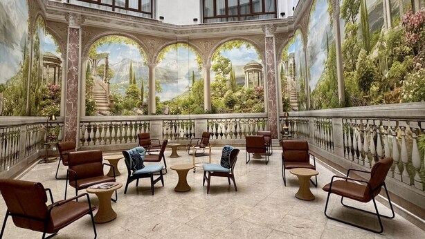 Een ruime binnentuin met een landschapsshildering, verschillende zithoeken met stoelen en tafels, en een balkon met een leuning.