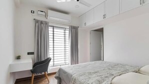A bedroom with a bed, a desk, and a chair. There is a window with curtains and an air conditioning unit on the wall.