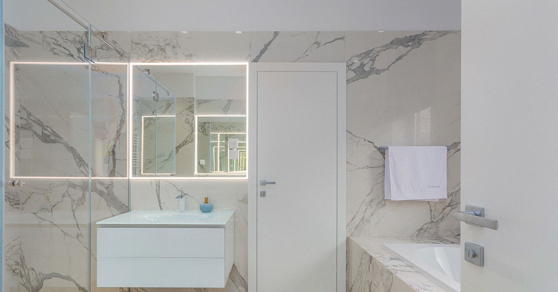 Une salle de bain moderne avec un pare-baignoire en marbre, une douche en verre et un meuble-lavabo blanc agrémenté d’un vase bleu.