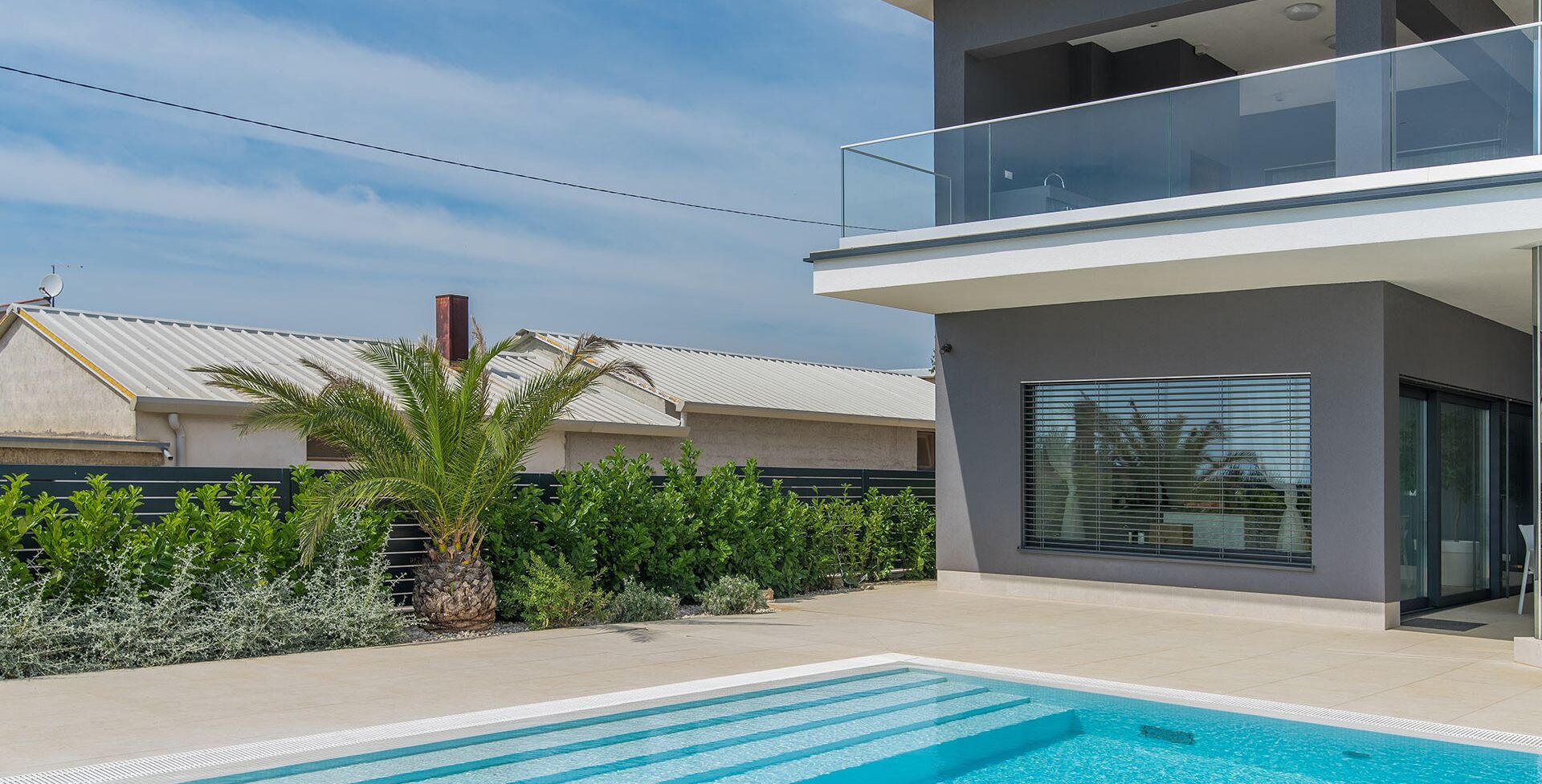A modern two-story building with a swimming pool, surrounded by lush greenery and a clear sky.
