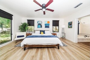Un dormitorio con cama, ventilador de techo, obras de arte, una planta y vistas al exterior.