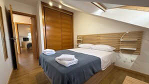 A hotel room with a wooden bed frame, a grey bedspread, white pillows, and a wooden headboard. There is a wooden wardrobe and a small wooden table.