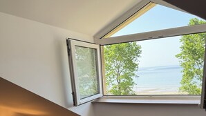 Ein Zimmer mit Meerblick durch ein großes Fenster, ein Stuhl mit gestreiftem Kissen und ein kleiner Tisch mit einer Lampe.