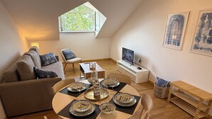 Ein Essbereich mit einem runden Tisch für vier Personen, einer Couch, einem Fernseher und einem Fenster mit Blick auf Grünflächen.
