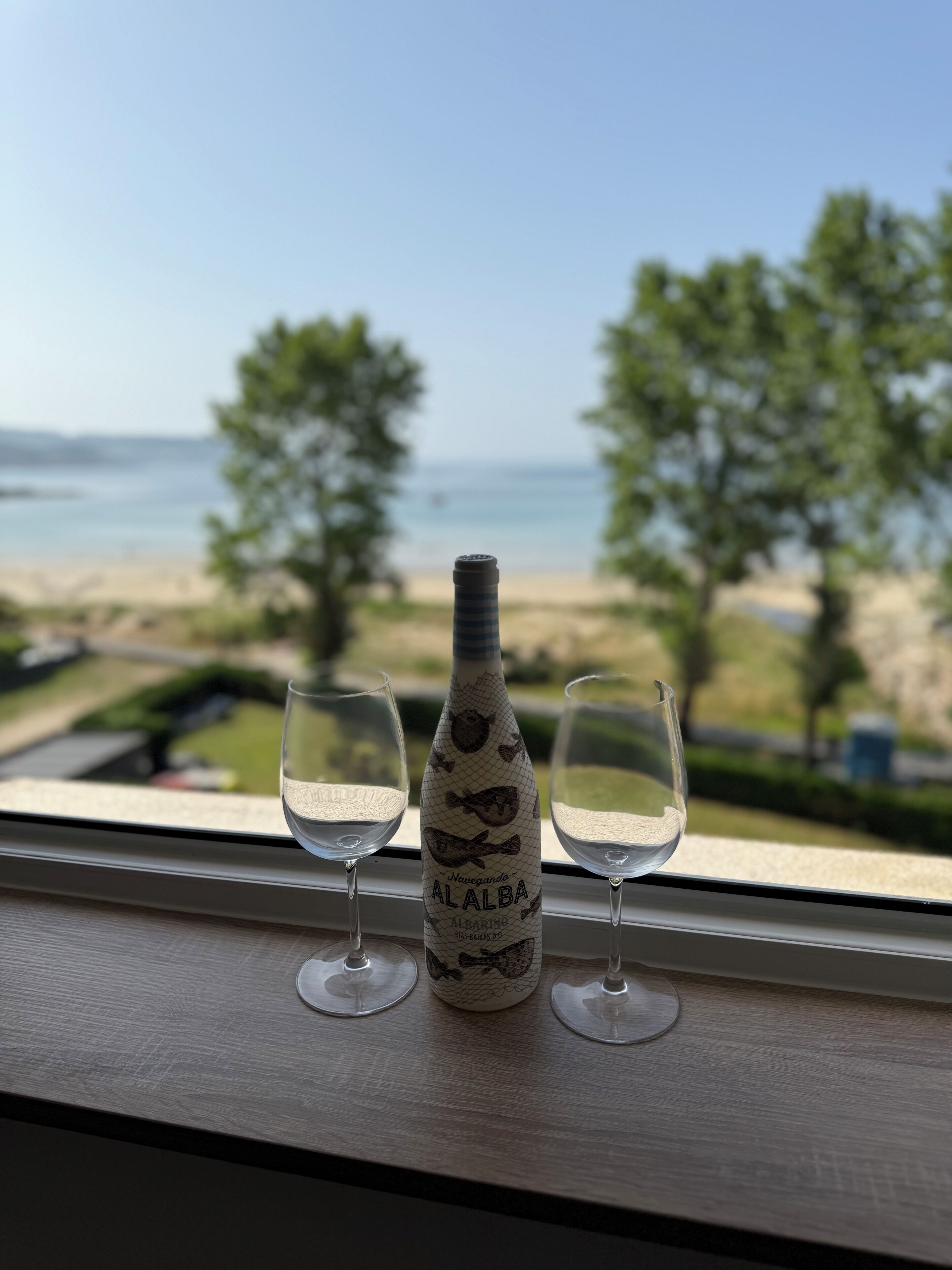 A bottle of Albarino wine and two glasses on a windowsill with a view of the beach.