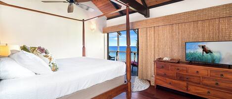 A bedroom with a wooden four-poster bed, a ceiling fan, a television on a wooden dresser, and a view of the ocean.