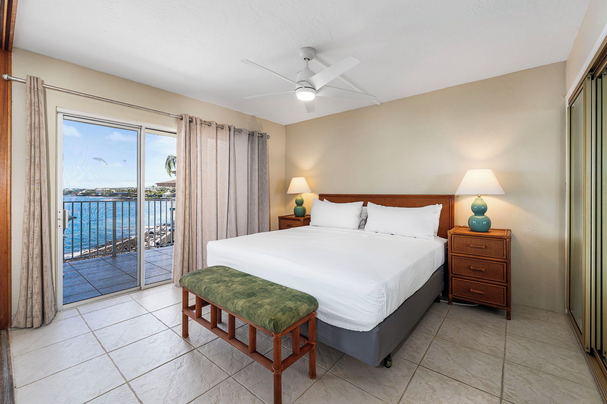 A hotel room with a large bed, two bedside tables with lamps, a bench, and a sliding glass door leading to a balcony with a view of the sea.