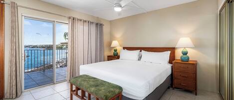A hotel room with a large bed, two bedside tables with lamps, a bench, and a sliding glass door leading to a balcony with a view of the sea.