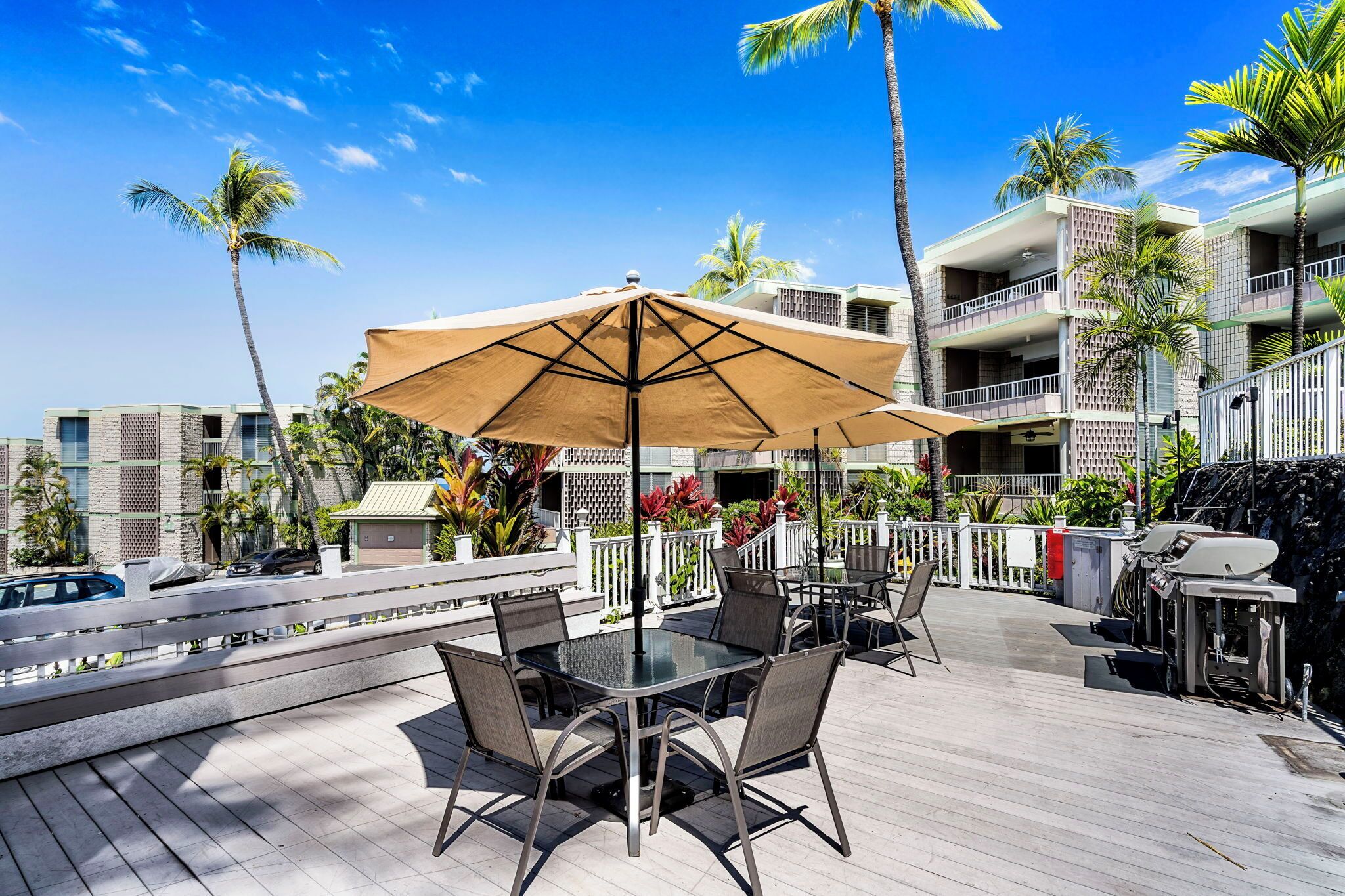 An outdoor patio with a table and chairs, a large umbrella, and a view of a building with balconies and palm trees.
