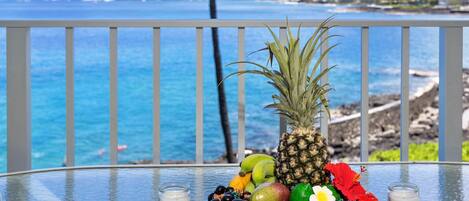A table with a tropical fruit arrangement and two glasses of milk on a balcony overlooking the ocean.