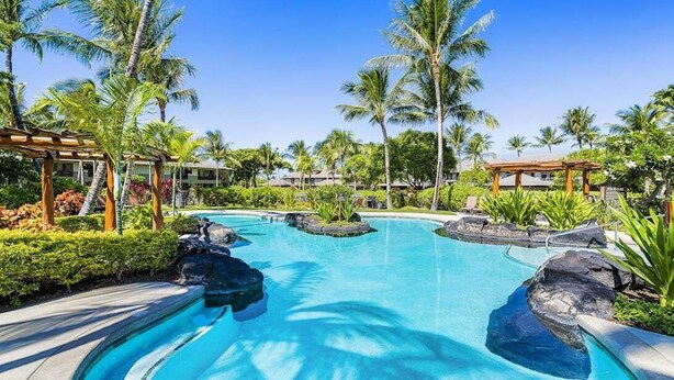 En pool på ett semesterhotell, omgiven av palmer, med en träpergola och en klarblå himmel.