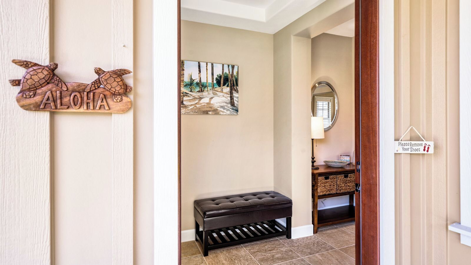 A hallway with a wooden sign reading "ALOHA," a black bench, a painting of a beach scene, a mirror, and a "Please Remove Your Shoes" sign.