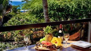 A table set with a pineapple, a bottle of wine, glasses, and tropical fruits, overlooking a lush garden and the ocean.
