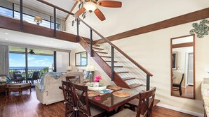 A spacious living area with a wooden staircase, a dining table with chairs, and a large window offering a view of the outdoors. - AC - Beautiful Ocean & Golf Views - 2 Bedrooms w/ Loft - Vrkcv (Kailua-Kona)
