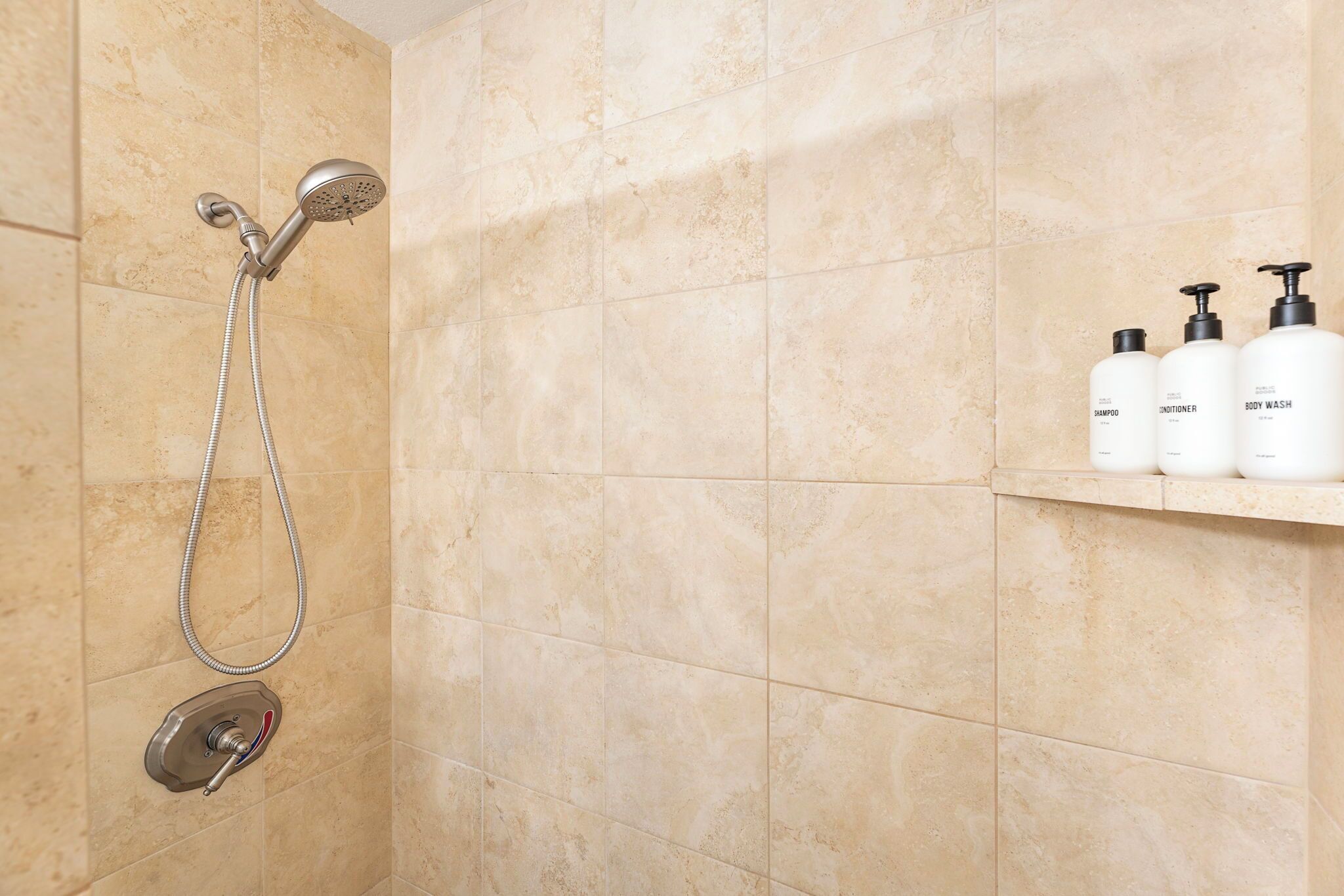 Un baño con baldosas color beige, una ducha con cabezal de ducha manual y un estante con tres botellas blancas etiquetadas como champú, acondicionador y gel de baño.