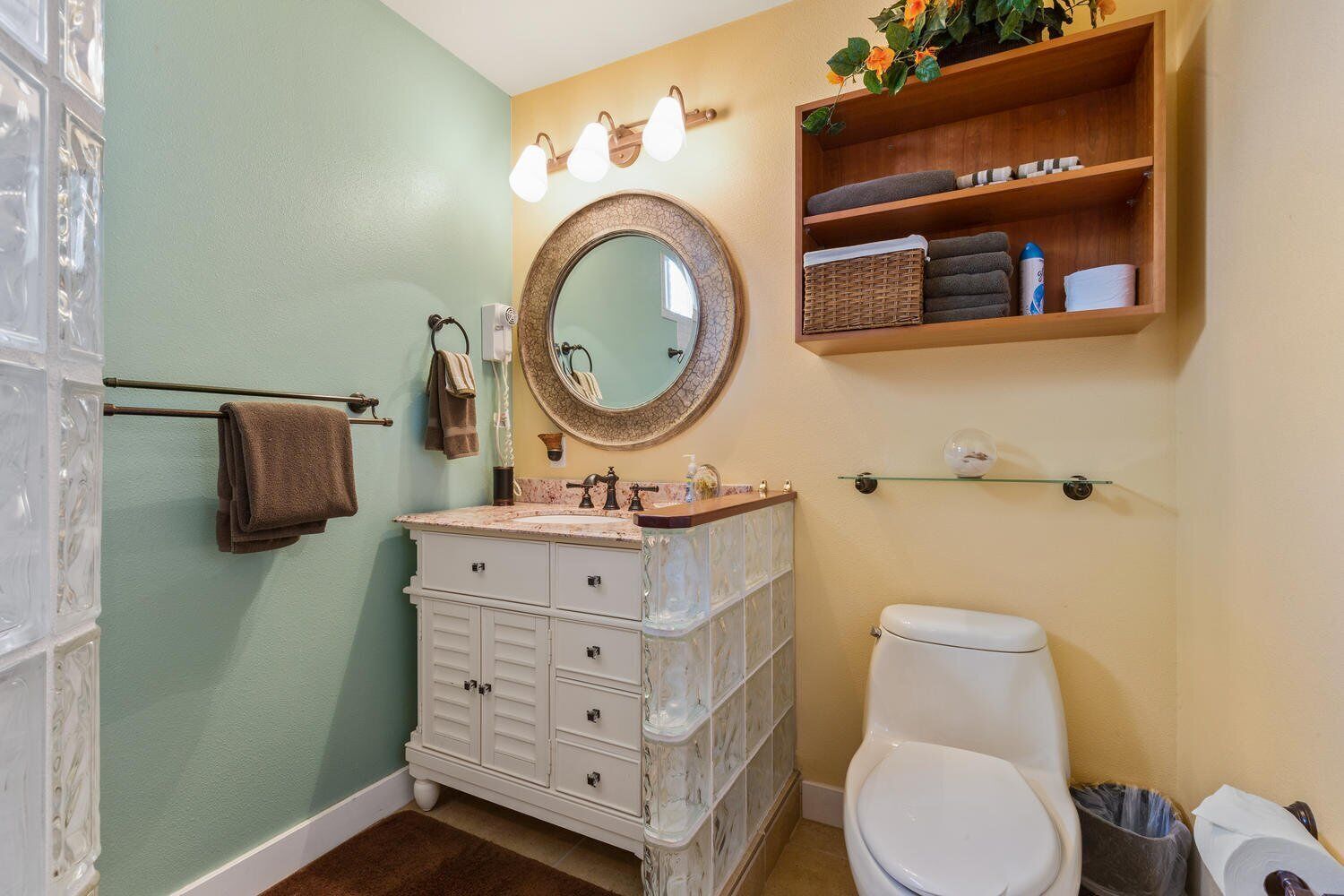 A bathroom with a white vanity, a round mirror, a toilet, and a shelf with towels and toiletries.
