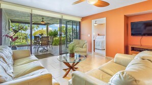 A spacious living area with a glass wall, a ceiling fan, a TV, and a dining area with outdoor views. - Ocean and Golf Course Views - Pool- vrKCV (Kailua-Kona)
