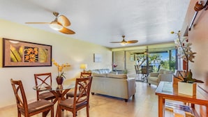 A spacious living area with a glass door leading to a balcony, a dining table with chairs, and two ceiling fans. - Ocean and Golf Course Views - Pool- vrKCV (Kailua-Kona)