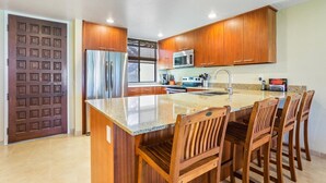 A kitchen with wooden cabinets, a stainless steel refrigerator, a microwave, and a granite countertop. - Ocean and Golf Course Views - Pool- vrKCV (Kailua-Kona)