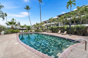 A swimming pool with a tiled edge, surrounded by palm trees and lounge chairs, with a building in the background. - Complete Renovation - 20 Steps to Ocean -washer/dryer in Unit- Vrkcv (Kailua-Kona)