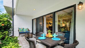 An outdoor dining area with wicker furniture, a table set with blue plates and fruit, and a bench with a colorful cushion. - Complete Renovation - 20 Steps to Ocean -washer/dryer in Unit- Vrkcv (Kailua-Kona)