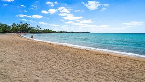 A beach with clear blue water, a sandy shore, and a few people walking.