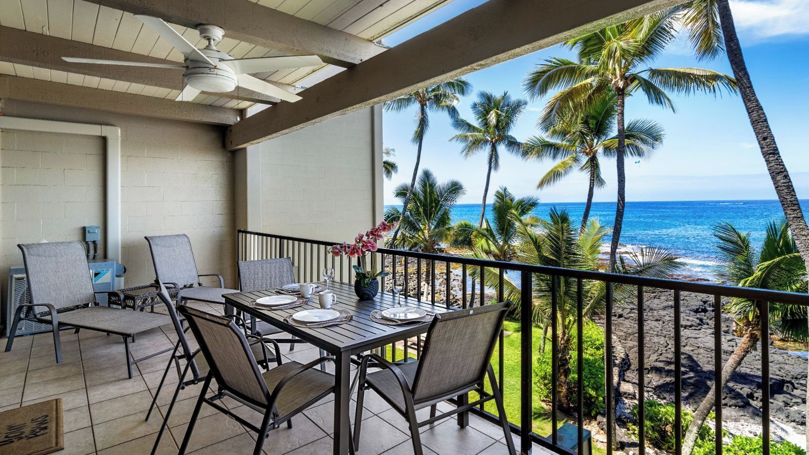 Un balcón con una mesa lista para comer, con vista a una playa con palmeras y el océano.