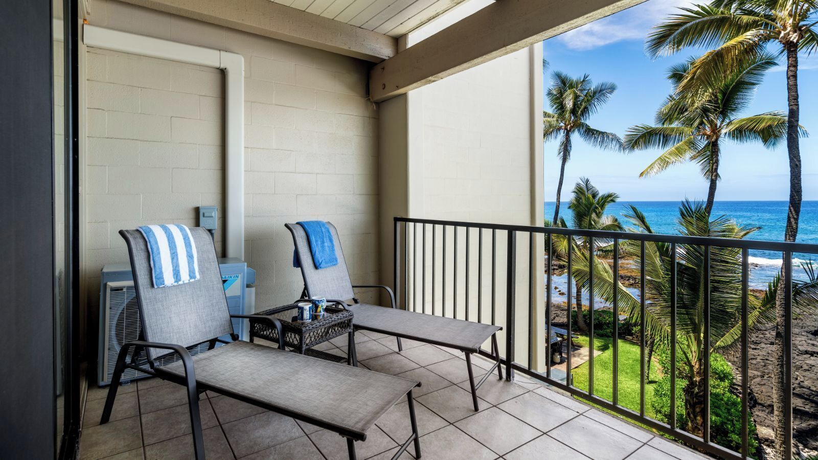 A balcony with two chairs and a table, overlooking a beach with palm trees and the ocean.