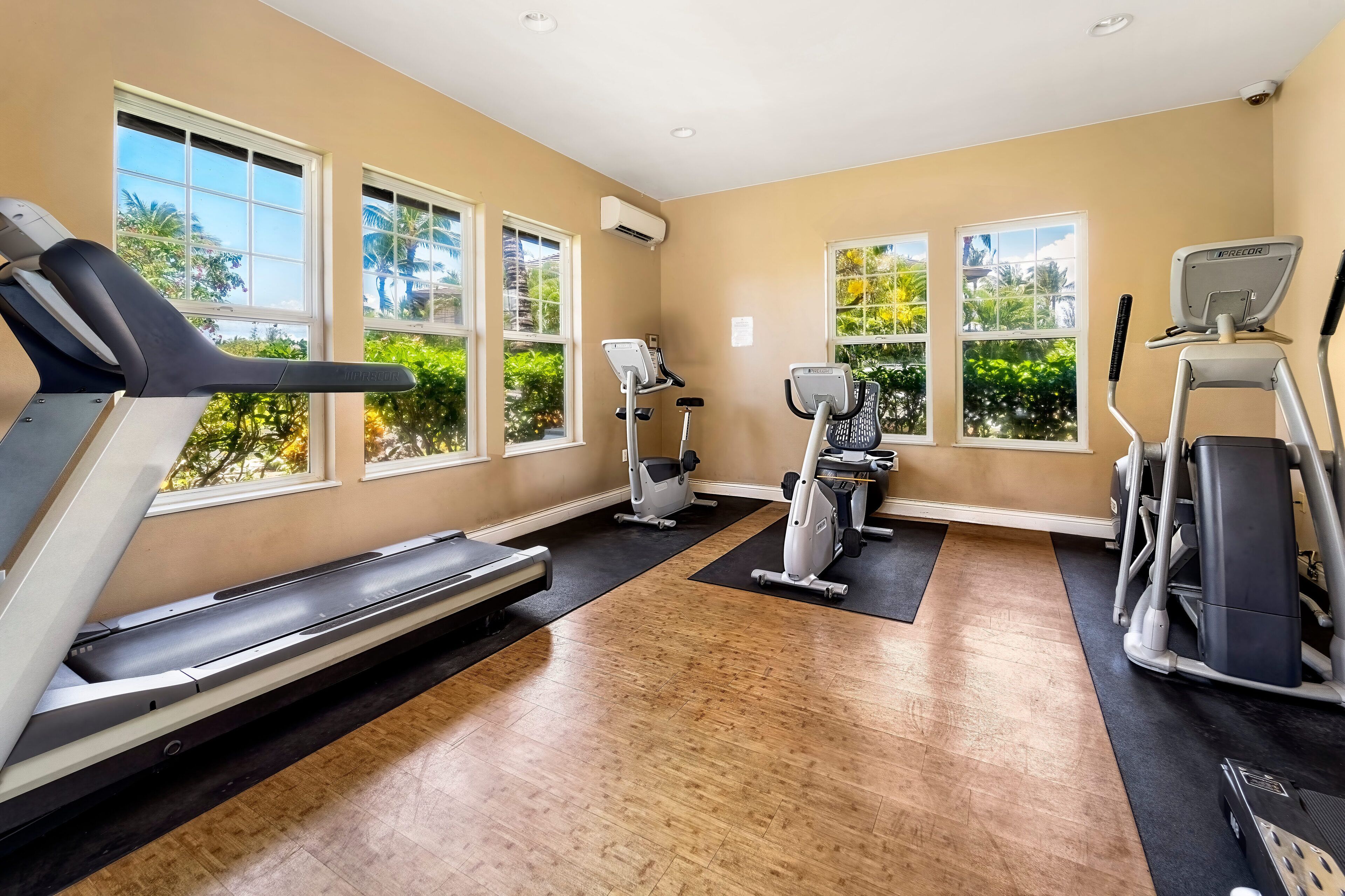 Un gimnasio con cintas de correr, elípticas y bicicletas estáticas, amplias ventanas con vistas a zonas verdes y suelo de madera.