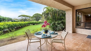 A patio with a round table set for two, surrounded by chairs, with a view of a well-maintained garden and a building in the background.