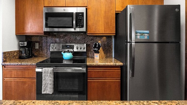 A kitchen with wooden cabinets, a stainless steel microwave, a black refrigerator, a stove with a blue kettle, and a coffee maker. - Great Location! - Ground Floor w/ AC - Beautiful Resort - Vrkcv (Waikoloa)