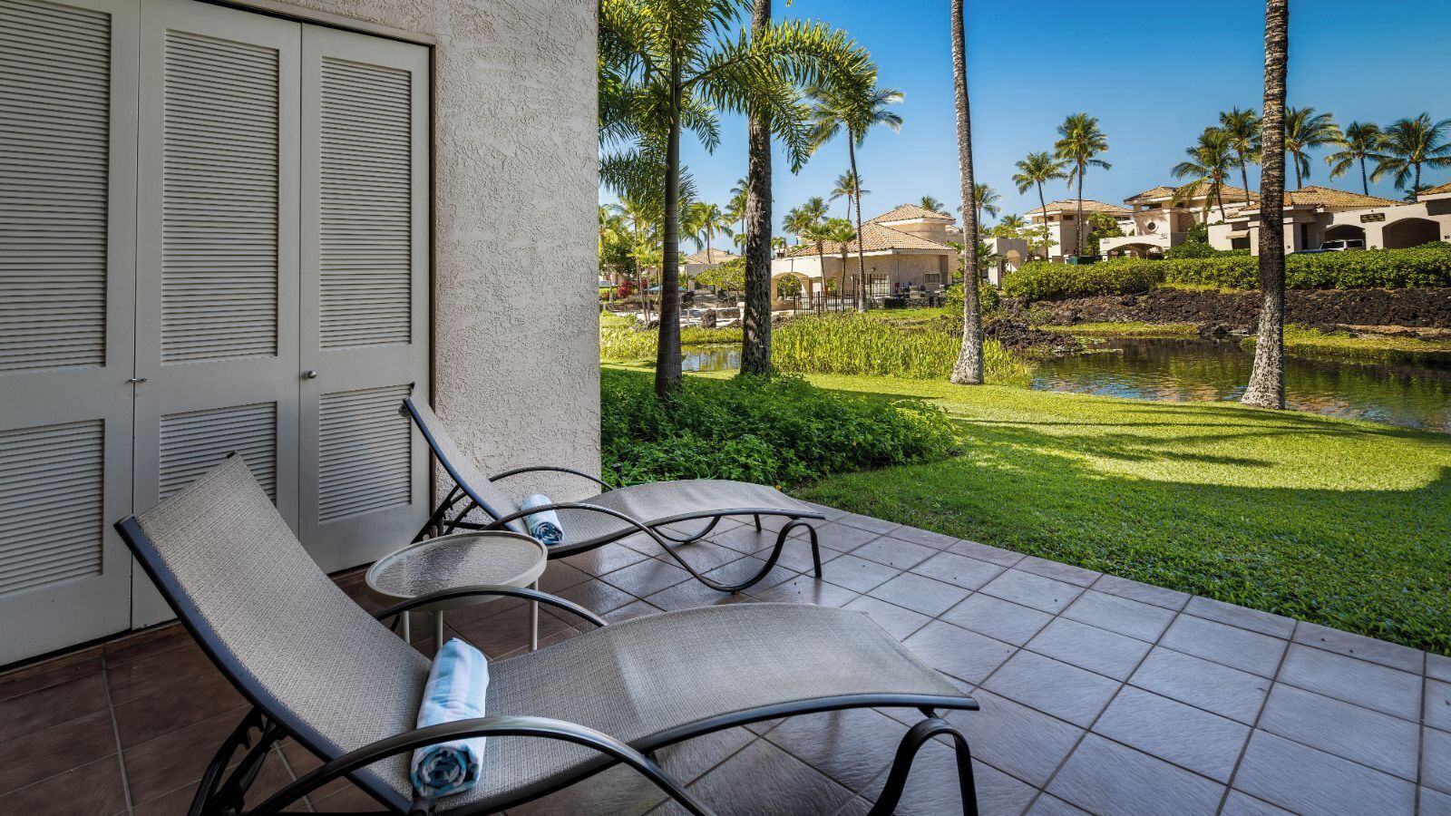 A patio with two lounge chairs and a small table, overlooking a pool and palm trees.