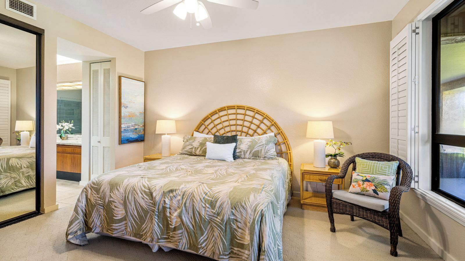 A bedroom with a large bed, a wicker chair, a nightstand with a lamp, and a window with blinds.