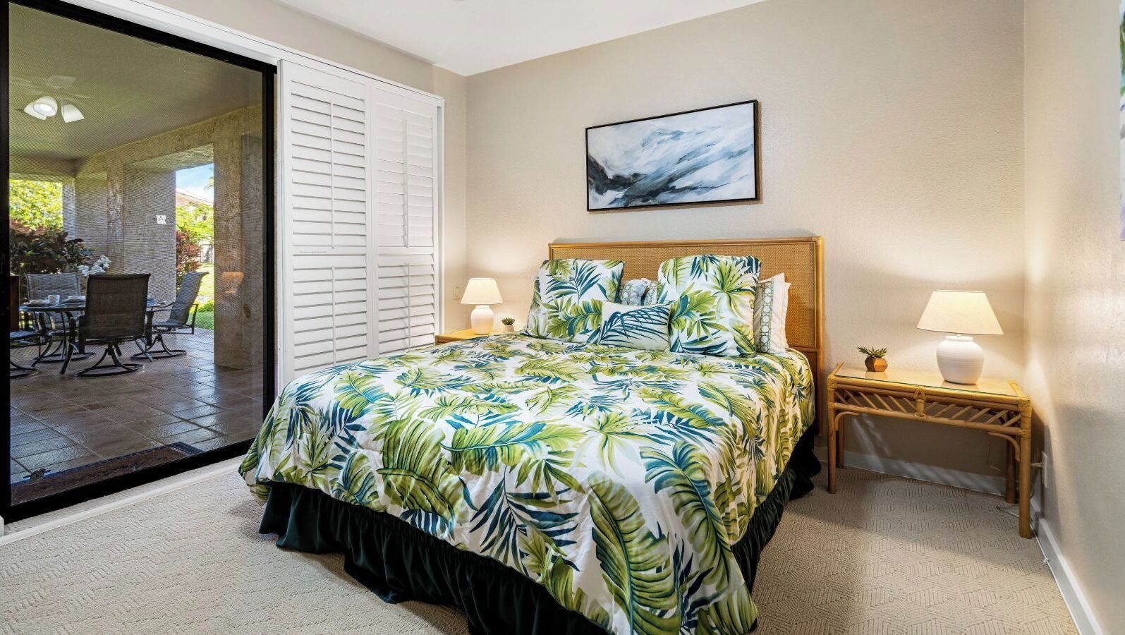 A bedroom with a bed, two bedside tables with lamps, a large window with blinds, and a view of an outdoor patio.